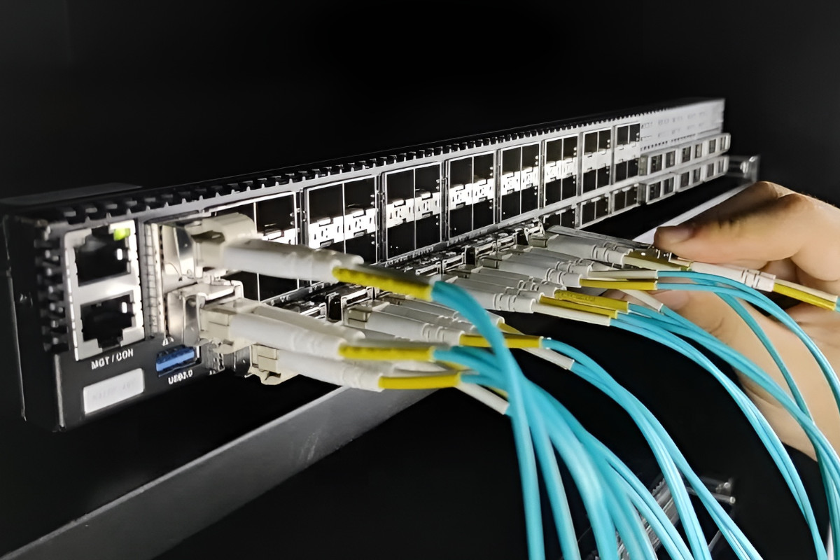 A hand connects multiple fiber optic cables to ports on a network switch in a server rack.