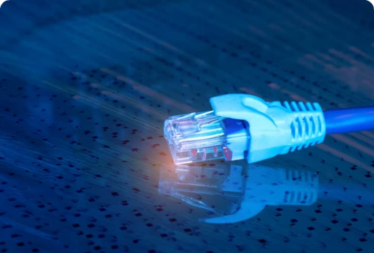 A close-up of a blue Ethernet cable with an RJ45 connector resting on a reflective surface, illuminated by blue light.